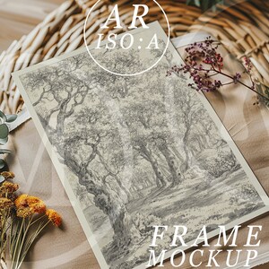 Op de afbeelding: Een ingelijste kunstdruk met een zwart-wit bossc&egrave;ne. Het kunstwerk wordt gepresenteerd op een beige stof, met gedroogde bloemen en een rieten mand op de achtergrond. De tekst "AR ISO:A" en "FRAME MOCKUP" zijn zichtbaar.