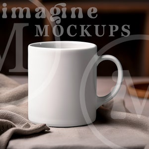 May include: A white ceramic mug with a curved handle sits on a neutral-colored cloth. The mug is the focal point, with the words "imagine MOCKUPS" subtly visible in the background. A lit candle adds warmth to the scene.
