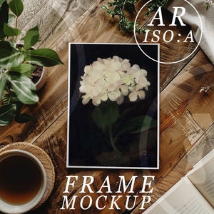 May include: A framed print of a white hydrangea flower on a black background. The frame is on a wooden table with a cup of coffee, a potted plant, and an open book.
