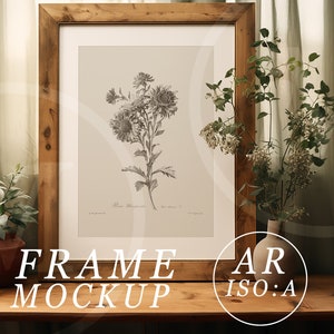 May include: A framed botanical print featuring a detailed black and white illustration of flowers. The artwork is set within a rustic, light brown wooden frame. The words "FRAME MOCKUP" are displayed at the bottom of the frame. The frame is on a wooden table with plants.