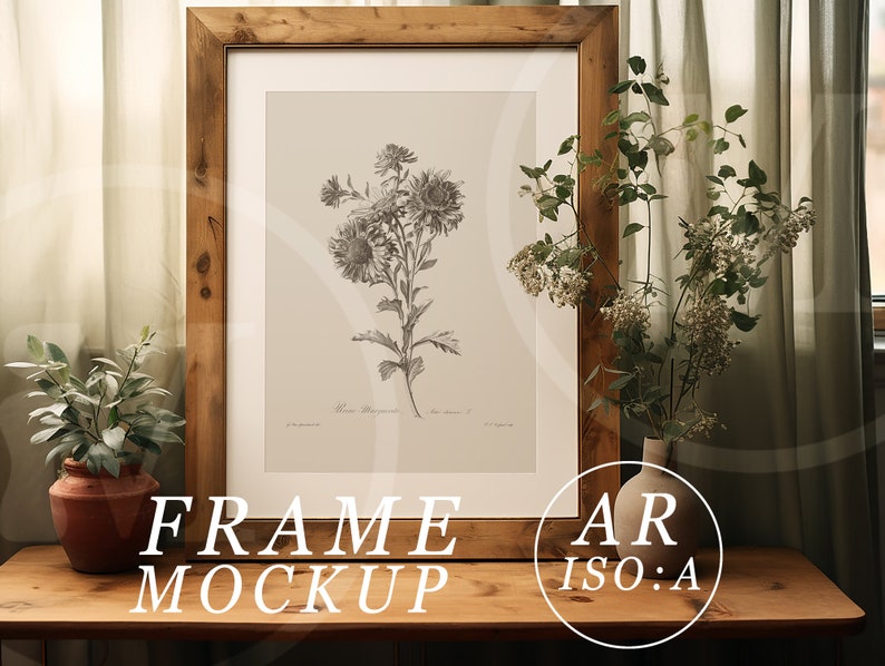 May include: A framed botanical print featuring a detailed black and white illustration of flowers. The artwork is set within a rustic, light brown wooden frame. The words "FRAME MOCKUP" are displayed at the bottom of the frame. The frame is on a wooden table with plants.