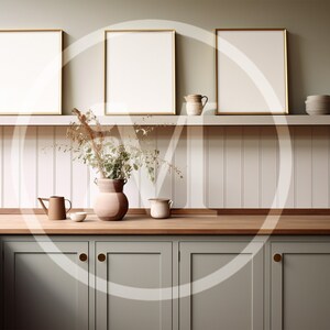 K&ouml;nnte beinhalten: K&uuml;cheninterieur mit drei gerahmten, leeren Kunstdrucken mit Goldrand &uuml;ber einer Arbeitsplatte. Die K&uuml;che hat salbeigr&uuml;ne Schr&auml;nke, eine Holz-Arbeitsplatte und ein Regal mit dekorativen Gegenst&auml;nden, darunter eine Vase mit Trockenblumen.