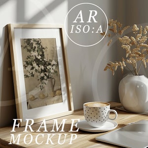 May include: A wooden frame with a white mount and a picture of a floral arrangement inside. The frame is sitting on a wooden table with a cup of coffee, a vase with dried flowers, and a laptop.