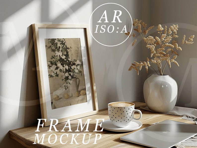 May include: A wooden frame with a white mount and a picture of a floral arrangement inside. The frame is sitting on a wooden table with a cup of coffee, a vase with dried flowers, and a laptop.