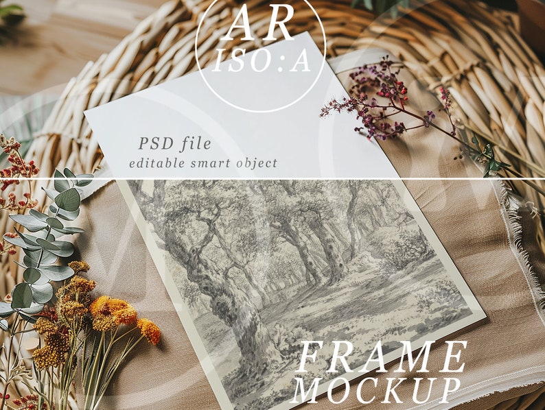 Op de afbeelding: Een frame-mockup met een zwart-wit landschapsafdruk met een bossc&egrave;ne, rustend op een beige stof. De afbeelding bevat gedroogde bloemen en de tekst "PSD file editable smart object" en "FRAME MOCKUP". Een gevlochten mand vormt de achtergrond.