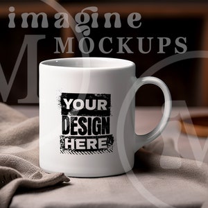 May include: A white ceramic mug with a curved handle. The mug features the text "YOUR DESIGN HERE" in a black, distressed font. The mug is set on a neutral-colored cloth, with a blurred background including a candle and the words "imagine MOCKUPS".