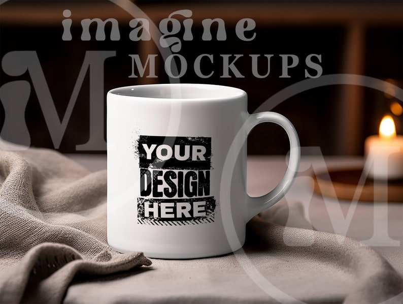 May include: A white ceramic mug with a curved handle. The mug features the text "YOUR DESIGN HERE" in a black, distressed font. The mug is set on a neutral-colored cloth, with a blurred background including a candle and the words "imagine MOCKUPS".