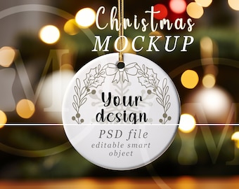 May include: A white ceramic Christmas ornament with a gold string, featuring a snowflake and floral design. The text "Your design" and "PSD file editable smart object" are printed on the ornament. The background is blurred with Christmas lights.