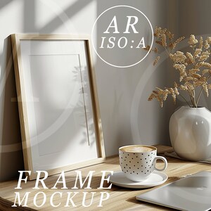 May include: A wooden frame mockup with a white mount and a white background. The frame is sitting on a wooden table with a cup of coffee, a vase with dried flowers, and a laptop.