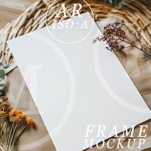 Op de afbeelding: Een witte frame mockup met de tekst "AR ISO:A" en "FRAME MOCKUP". Het frame ligt op een beige stof, omringd door gedroogde bloemen en een rieten mand.