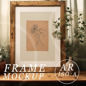 May include: A framed sunflower sketch in a rustic wooden frame. The artwork is on a tan background with a white mat. The words "FRAME MOCKUP" are at the bottom of the frame. A small plant in a terracotta pot and a vase of flowers are on the wooden table.