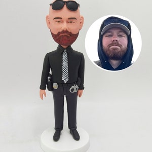 May include: A custom bobblehead figurine of a person wearing a black shirt, black pants, a black tie, and sunglasses. The figurine is holding a badge and handcuffs. The person's face is visible and has a beard.