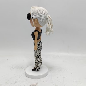 May include: A white figurine of a woman with blonde hair wearing black sunglasses, a black crop top, and black and white patterned pants. The figurine is standing on a white base.
