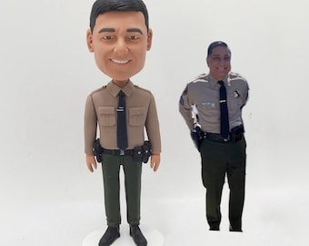 May include: A custom bobblehead figurine of a person wearing a tan uniform with a black tie, black belt, and green pants. The figurine is standing on a white base. The person is smiling and has black hair. A photo of the person in a similar uniform is visible in the background.