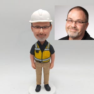 Peut inclure: Une figurine personnalisée d'une personne portant un casque blanc, une chemise noire, un gilet de sécurité jaune, un pantalon kaki et des chaussures noires. La figurine est debout sur une base blanche.