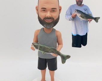 May include: A custom bobblehead figurine of a person wearing a gray tank top and black shorts, holding a green and brown fish. The figurine is standing on a white base. The person has a beard and brown hair.