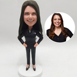 Op de afbeelding: Een gepersonaliseerd bobblehead-beeldje van een vrouw met lang bruin haar, gekleed in een zwart jack en een zwarte broek. Het beeldje staat op een witte voet.