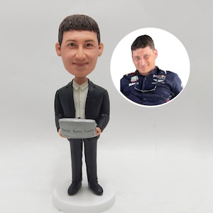 Op de afbeelding: Een gepersonaliseerd bobblehead-beeldje van een man in een zwart pak die een kleine grijze steen vasthoudt met de tekst "Best Boss Ever" erop. Op de achtergrond is een kleinere foto van de man te zien.