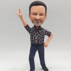 May include: A figurine of a man with dark hair and a beard, wearing a black shirt with a red and white floral pattern, blue pants, and black shoes. The figurine is on a white base with the text "#1 MMH IM Program Manager".