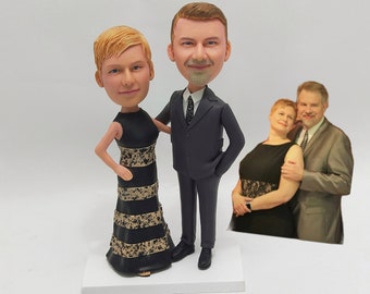 May include: A custom-made figurine of a couple dressed in formal attire. The woman is wearing a black and gold dress, and the man is wearing a black suit with a white shirt and a tie. The figurines are standing on a white base.