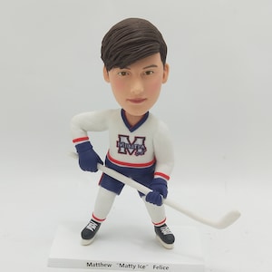 Op de afbeelding: Een gepersonaliseerde bobblehead van een hockeyer die een wit shirt draagt met een blauwe en rode "M" voor "Militia" op de voorkant. De speler houdt een hockeystick vast en staat in een schaatspose. De basis van de bobblehead heeft de naam "Matthew "Matty Ice" Felice" erop.