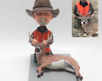 May include: A custom bobblehead figurine depicting a person wearing a brown hat, a bright orange vest, and camouflage clothing, kneeling behind a brown deer with antlers. The figurine is mounted on a rectangular gray base, accompanied by a reference photo.