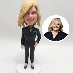 May include: A custom bobblehead figurine of a woman with blonde hair wearing a black jacket and black pants. The figurine is standing on a white base. A photo of the woman is shown in a circle behind the figurine.