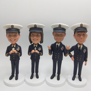 Può includere: Quattro statuette a testa oscillante di persone in uniforme della marina con cappelli bianchi. Ogni statuetta è su una base circolare bianca. Le uniformi hanno dettagli dorati e medagliette in miniatura. Le statuette hanno volti sorridenti.