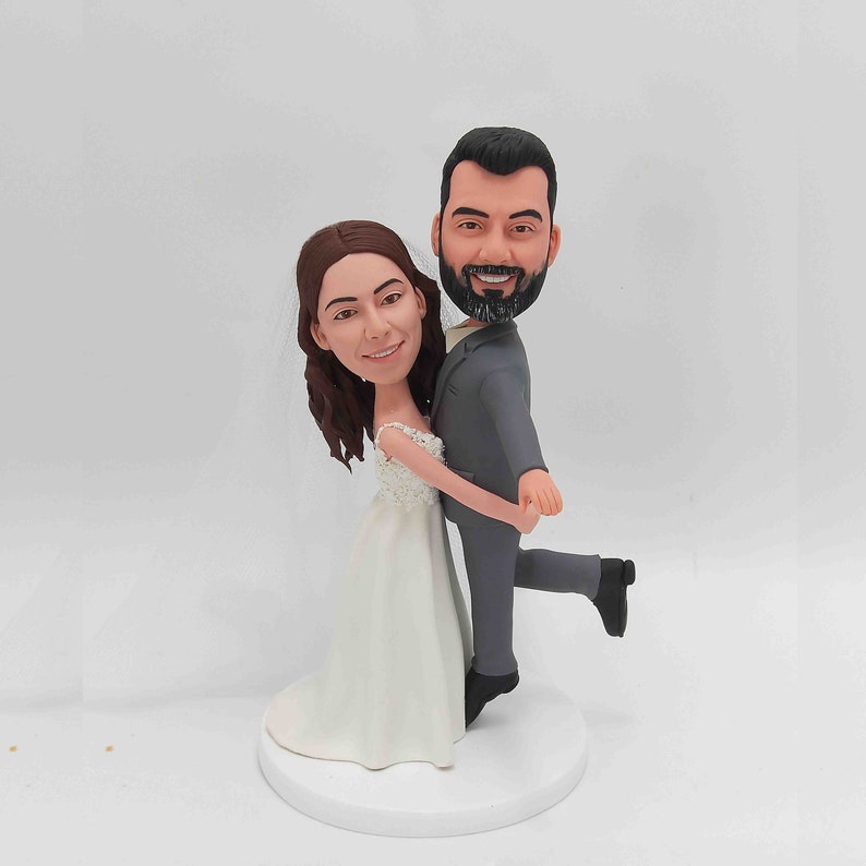 K&ouml;nnte beinhalten: Ein Hochzeitstortenaufsatz mit einer Braut und einem Br&auml;utigam in einer verspielten Pose. Die Braut tr&auml;gt ein wei&szlig;es Kleid und der Br&auml;utigam einen grauen Anzug. Der Br&auml;utigam h&auml;lt die Braut in seinen Armen und beide l&auml;cheln.
