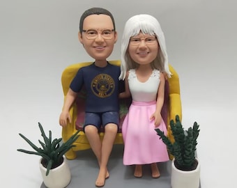 May include: A figurine of a couple sitting on a yellow couch. The man wears a navy blue shirt and shorts. The woman wears a white top and pink skirt. Two small potted plants flank the couch on a gray base.