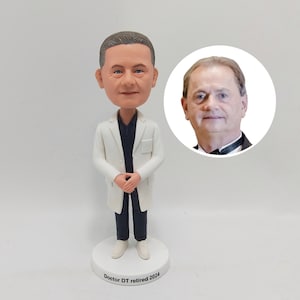 May include: A white doctor bobblehead figurine with a black shirt and a white lab coat. The bobblehead has a realistic face and is standing on a white base with the text "Doctor DT retired 2024".