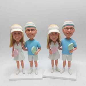 May include: Four bobblehead figurines of a couple, each holding a paddle. The woman wears a white visor and dress, while the man wears a blue shirt and shorts. The figurines stand on white bases, ideal for a personalized gift.