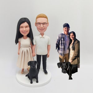 May include: A custom-made cake topper featuring a couple and their dog. The couple is depicted as cartoon figures with realistic features. The man is wearing a white polo shirt and black pants. The woman is wearing a white dress. The dog is black and sitting in front of the couple. The cake topper is on a white base.
