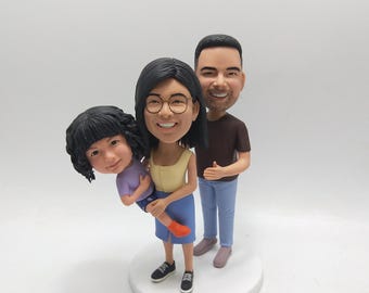 May include: A family figurine featuring a woman in a yellow top and blue skirt holding a child in a purple shirt. A man in a brown shirt and blue jeans stands behind them. The figures are on a white base.