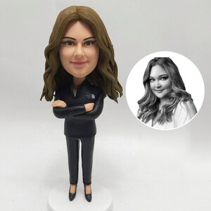 May include: A custom bobblehead figurine of a woman with brown hair, wearing a black jacket and pants. The figurine has a small base and a black and white photo of the woman is in the background.