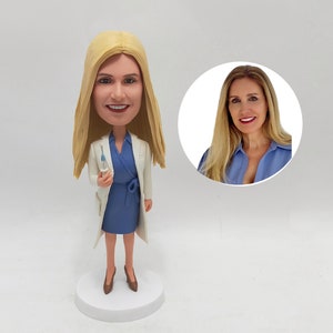 May include: A custom bobblehead figurine of a woman with blonde hair wearing a white lab coat and a blue dress. She is holding a syringe in her right hand. The figurine is standing on a white base.