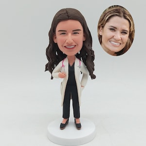 May include: A figurine of a female doctor with brown hair, wearing a white lab coat, black pants, and black shoes. A pink stethoscope hangs around her neck. An oval image of a smiling woman is in the background.