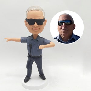 May include: A custom bobblehead figurine of a person wearing a grey uniform, black sunglasses, and a black belt. The figurine is standing with one arm outstretched and the other hand on their hip. The figurine is on a white base.