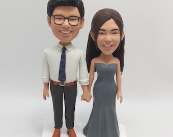 May include: A custom-made figurine set of a couple, the man is wearing a white shirt, a blue tie, black pants, and brown shoes. The woman is wearing a gray strapless dress and gray shoes. The figurines are holding hands.