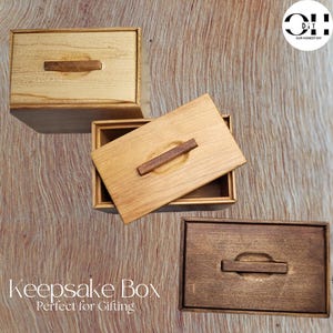 Puede incluir: Tres cajas de recuerdo de madera con tapas. Las cajas están hechas de diferentes tipos de madera y tienen un acabado natural. Cada caja tiene una manija de madera en la tapa. El texto "Keepsake Box Perfect for Gifting" está impreso debajo de las cajas.