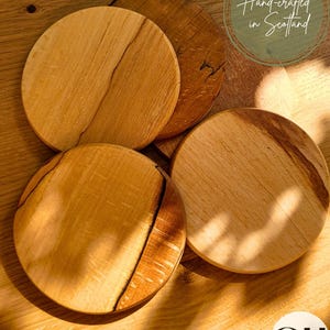 Puede incluir: Cuatro posavasos redondos de madera clara con vetas naturales y detalles marrón oscuro. Los posavasos están colocados sobre una superficie de madera clara. El texto "Hand-crafted in Scotland" es visible.