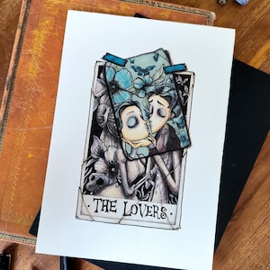 Peut inclure: Illustration à l'aquarelle d'une carte de tarot intitulée "Les Amoureux". La carte représente deux personnages s'embrassant, entourés de fleurs bleues et de papillons. L'œuvre est sur papier blanc, avec un livre vintage et du papier noir en arrière-plan.