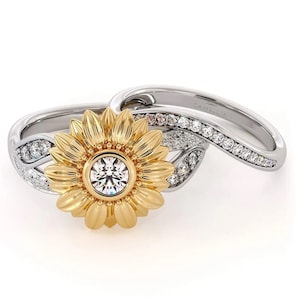 May include: A two-piece ring set featuring a sunflower design with a clear gemstone in the center. The sunflower is made of yellow gold and the band is made of white gold with small clear gemstones.