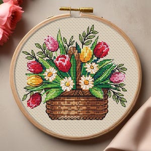 Puede incluir: Un bordado de punto de cruz de una cesta llena de flores coloridas, incluyendo tulipanes rojos, rosas y amarillos, y margaritas blancas. El bordado está enmarcado en un aro de madera, creando un diseño floral.