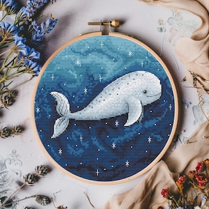 May include: A white whale with a blue tail swims through a dark blue starry sky. The whale is embroidered on a round canvas with a wooden frame.