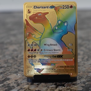 May include: A gold Charizard GX Pokémon card, featuring a colorful dragon illustration. The card displays the text "Charizard GX" and "HP250", along with attack details. The card is displayed on a black stand.