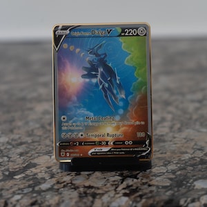 Pode incluir: Um cartão Pokémon Origin Forme Dialga V com borda dourada, exibido em um suporte preto. O cartão apresenta uma ilustração azul e branca de uma criatura em um fundo colorido. O texto inclui "Metal Coating" e "Temporal Rupture."