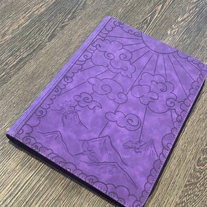 K&ouml;nnte beinhalten: Ein lila Notizbuch mit einem Sonnen-, Wolken- und Bergdesign. Der Einband ist mit einer detaillierten Illustration gepr&auml;gt. Das Notizbuch hat einen gen&auml;hten Rand und ein dunkelviolettes Innere.