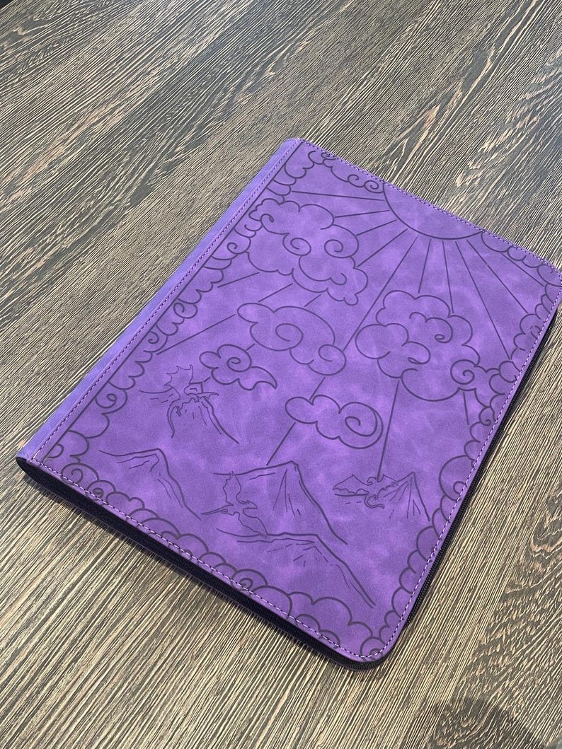 K&ouml;nnte beinhalten: Ein lila Notizbuch mit einem Sonnen-, Wolken- und Bergdesign. Der Einband ist mit einer detaillierten Illustration gepr&auml;gt. Das Notizbuch hat einen gen&auml;hten Rand und ein dunkelviolettes Innere.