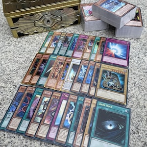 May include: A gold-coloured metal Yu-Gi-Oh! storage tin with an embossed eye design, accompanied by three stacks of trading cards and a collection of individual cards laid out in rows, including Dark Magician and Dark Hole spell cards.